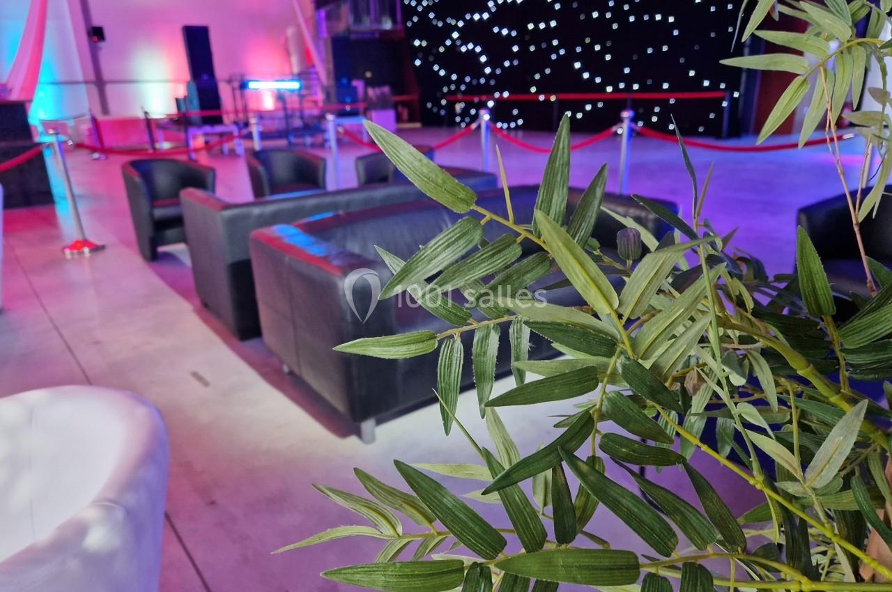 Plantes en premier plan avec des fauteuils noirs et un espace événementiel éclairé en arrière-plan.