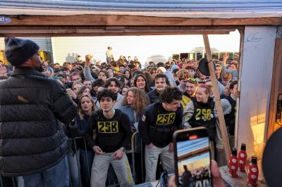 Un grand groupe de jeunes assis dehors sur un sol en béton, près de bâtiments sous un ciel nuageux.
