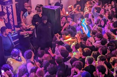 Un grand groupe de jeunes assis dehors sur un sol en béton, près de bâtiments sous un ciel nuageux.