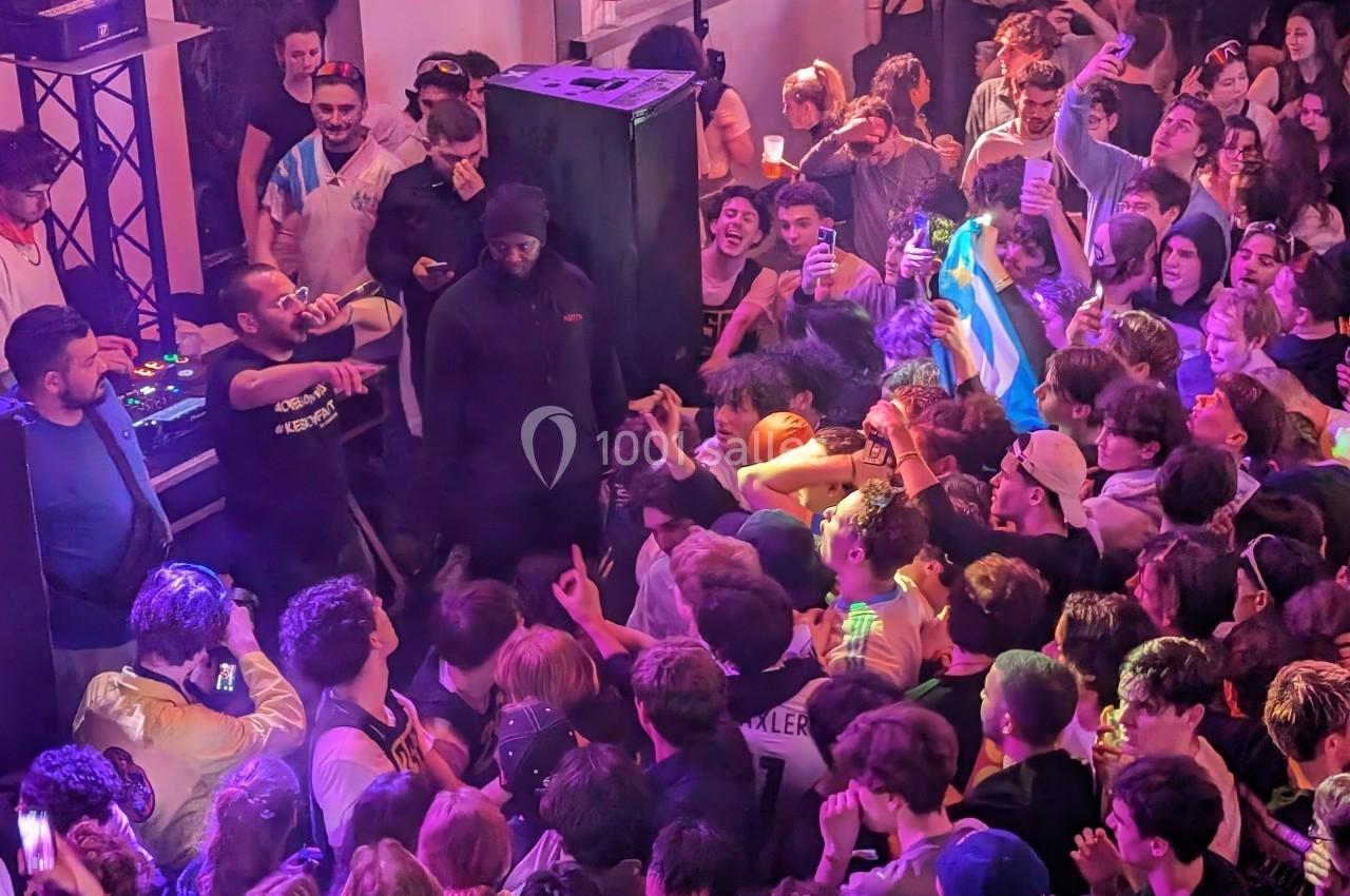 Foule rassemblée dans une salle de concert, avec des artistes sur scène et des spectateurs animés sous des lumières colorées.