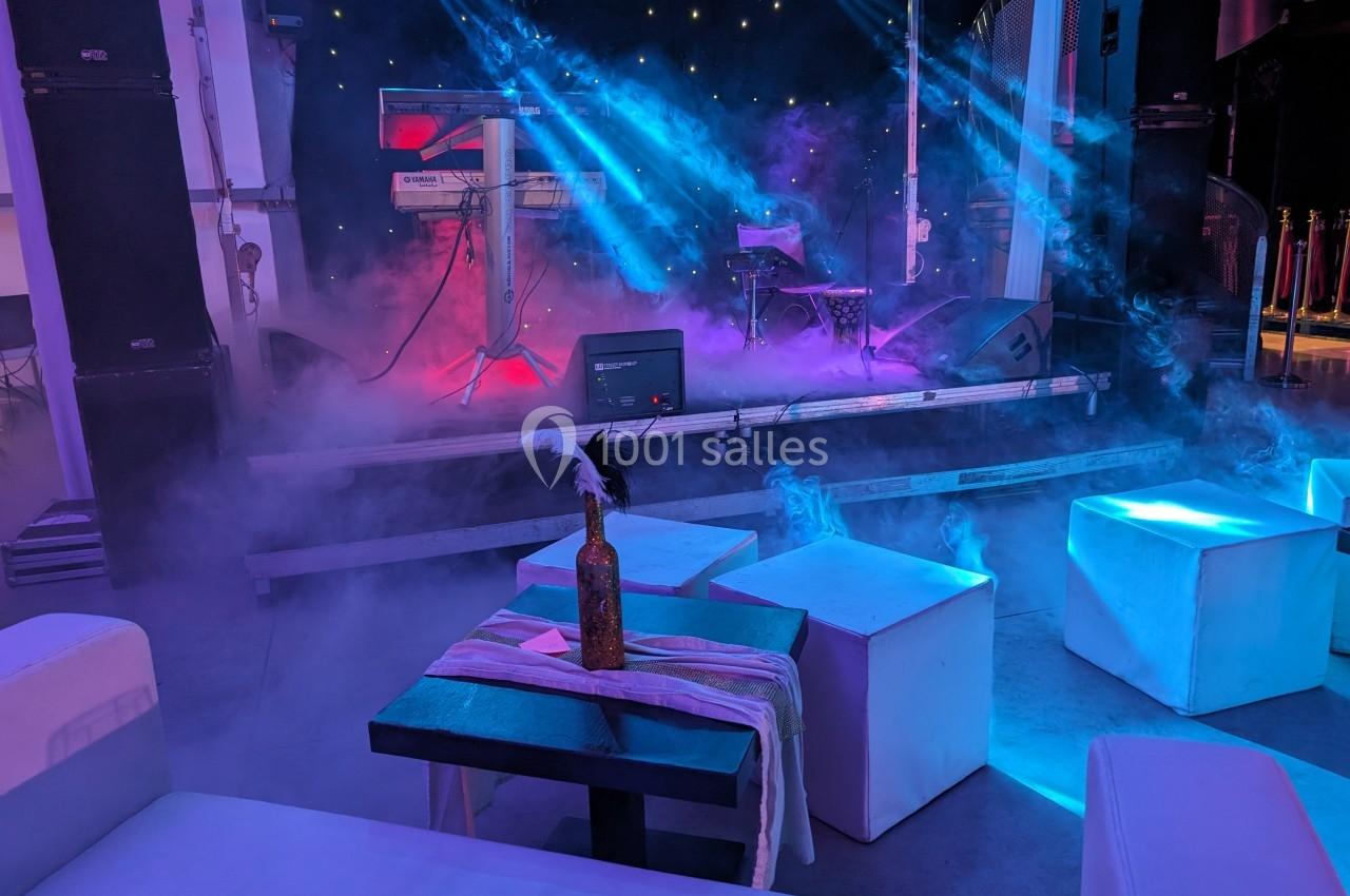 Scène éclairée avec des projecteurs colorés, fumée diffuse et tables basses entourées de poufs blancs.