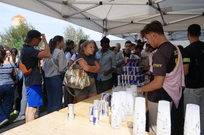 Un grand groupe de jeunes assis dehors sur un sol en béton, près de bâtiments sous un ciel nuageux.