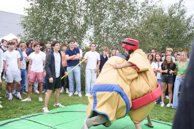 Un grand groupe de jeunes assis dehors sur un sol en béton, près de bâtiments sous un ciel nuageux.
