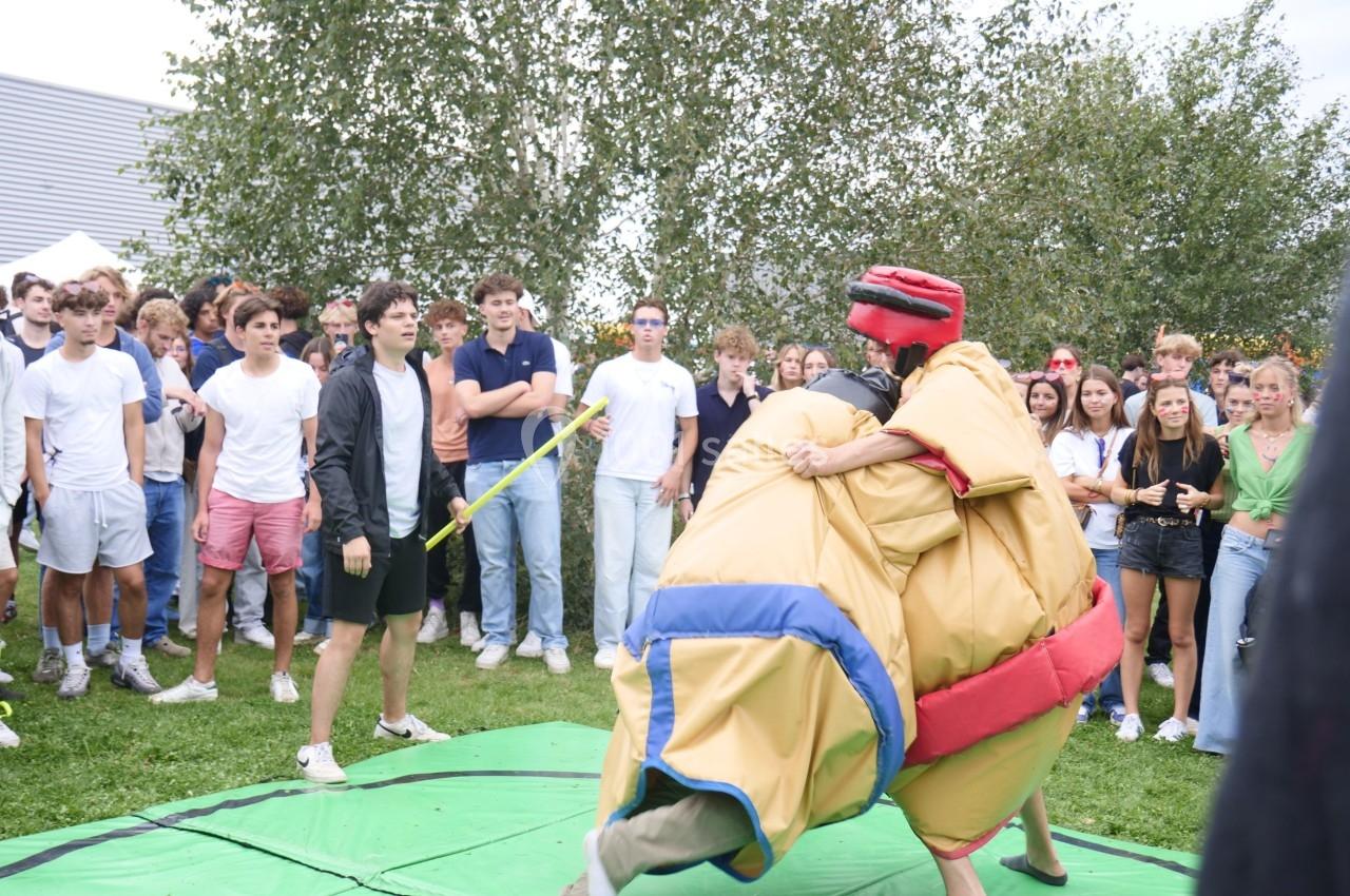 Deux personnes en costumes de sumo s'affrontent sur un tapis vert, entourées d'un groupe de spectateurs en extérieur.