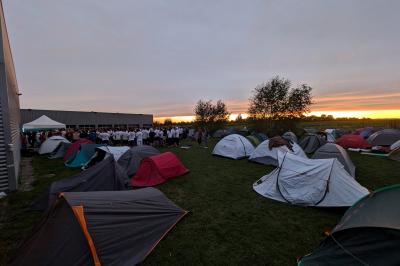 Un grand groupe de jeunes assis dehors sur un sol en béton, près de bâtiments sous un ciel nuageux.