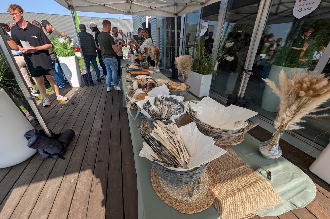 Buffet en extérieur sous un barnum, avec des paniers d'ustensiles, des plats variés et des invités en arrière-plan.