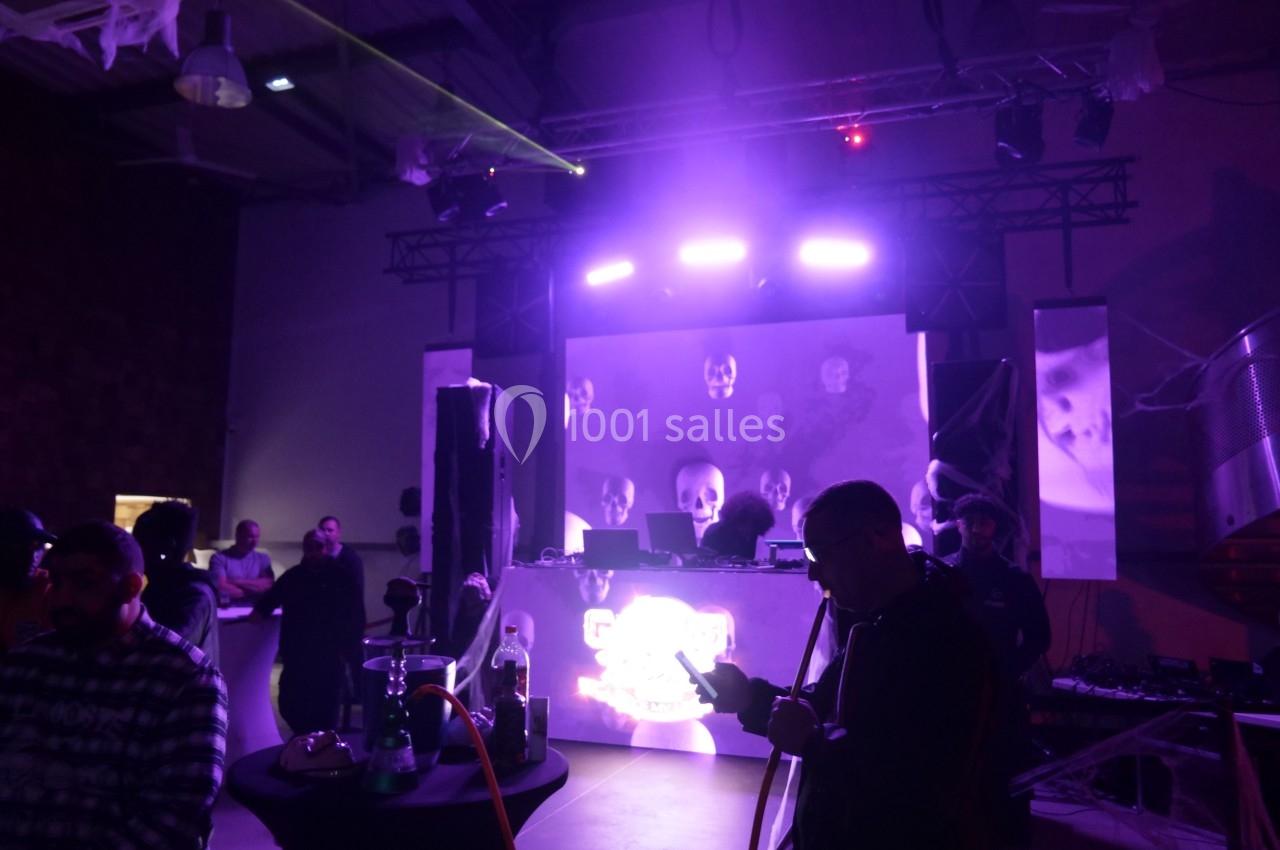 Salle sombre avec une scène éclairée par des lumières violettes, un DJ en arrière-plan et des personnes debout autour de…
