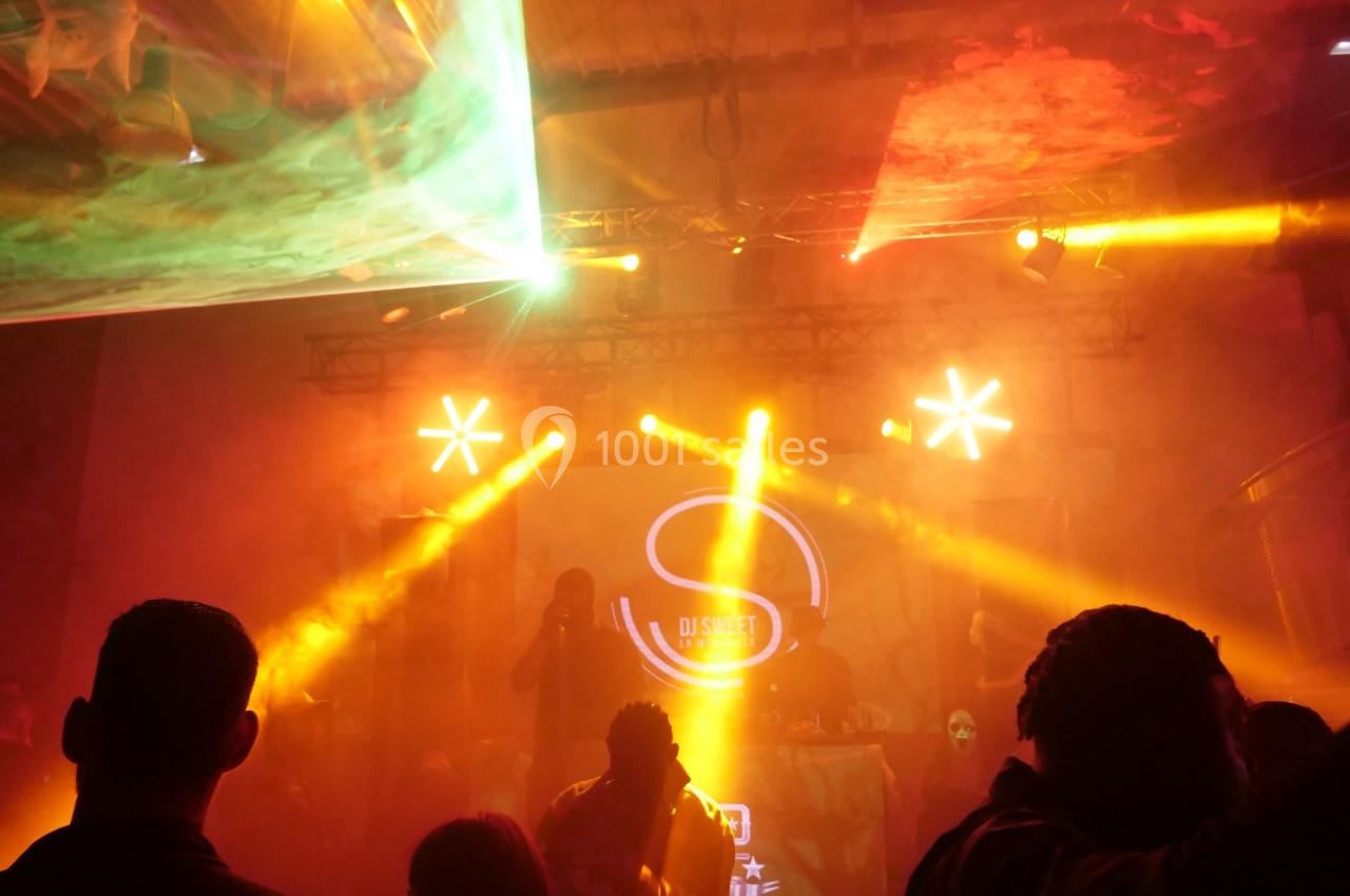 Salle de concert sombre avec éclairages colorés, une scène et des silhouettes de spectateurs au premier plan.