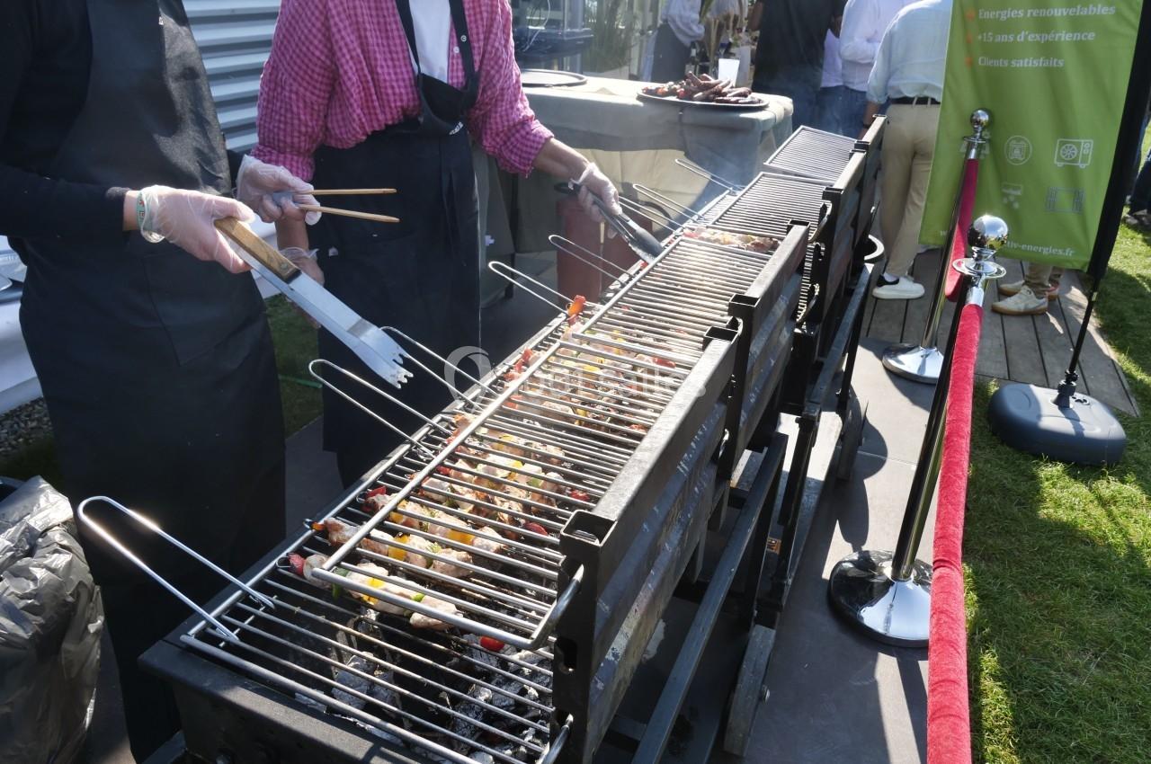 Deux personnes grillent des brochettes de légumes et de viande sur un grand barbecue lors d'un événement en extérieur.