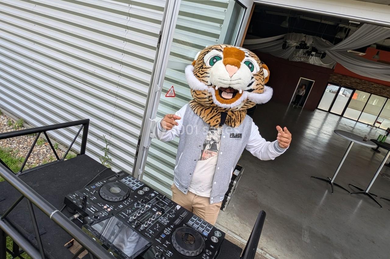 Personne portant une tête de tigre en peluche, debout derrière une table de mixage à l'extérieur d'un bâtiment.
