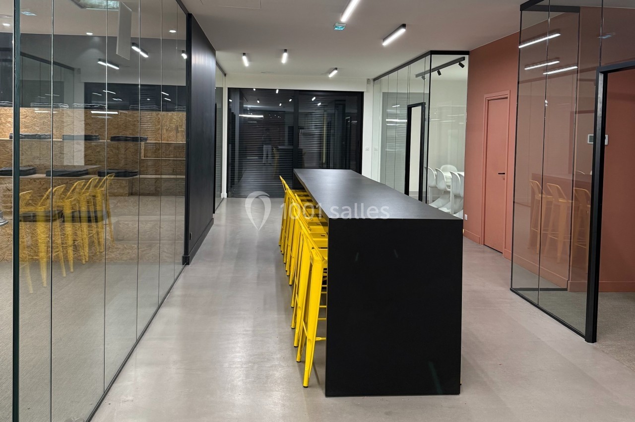 Salle moderne avec une longue table noire entourée de tabourets jaunes, séparée par des cloisons vitrées.