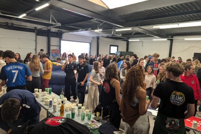 Un grand groupe de jeunes assis dehors sur un sol en béton, près de bâtiments sous un ciel nuageux.