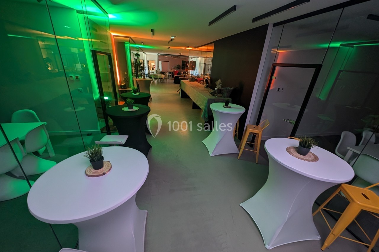 Salle moderne avec tables hautes blanches, chaises et éclairage coloré vert et orange, prête pour un événement.