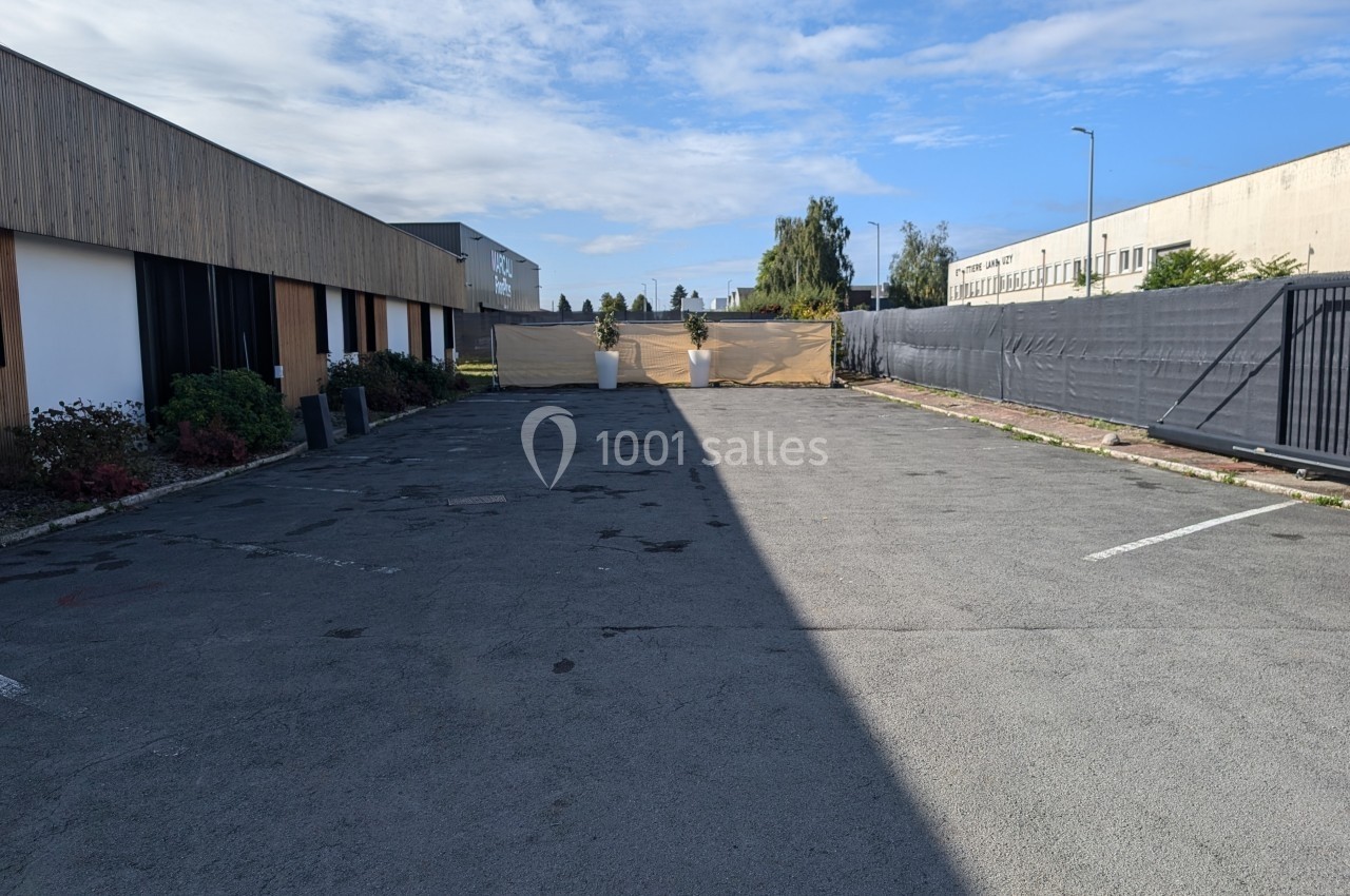 Parking vide entouré de bâtiments modernes, avec une clôture beige au fond et un ciel partiellement nuageux.