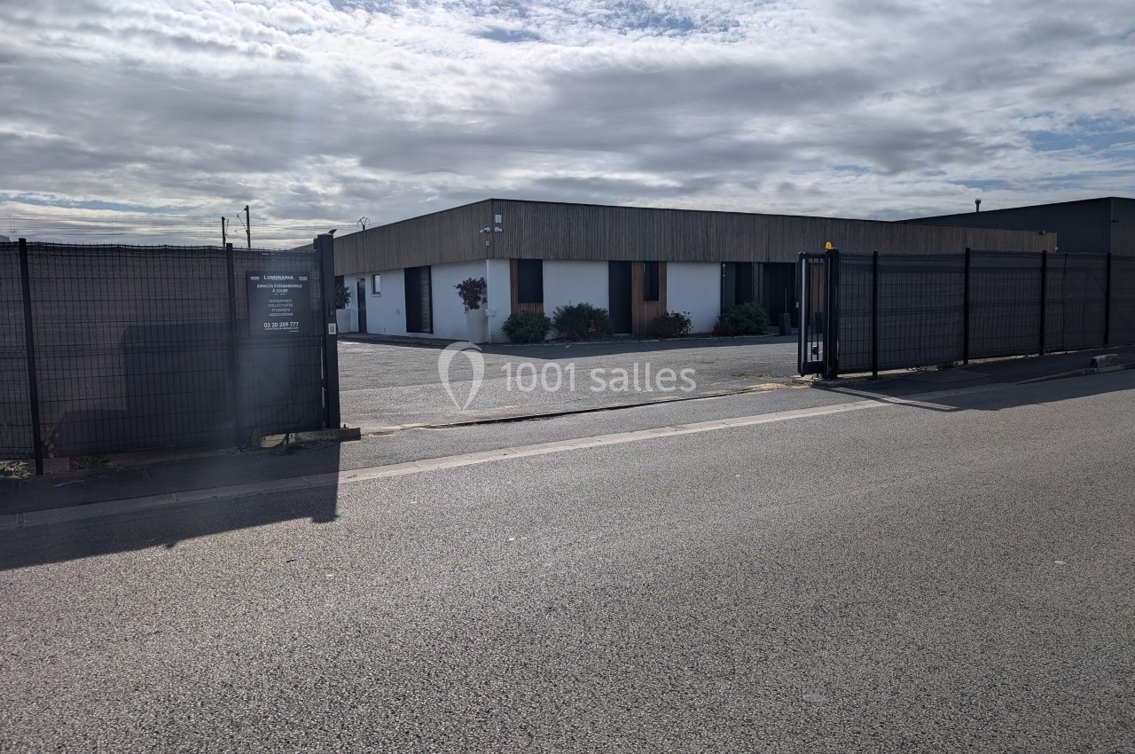 Bâtiment industriel entouré d'une clôture noire avec un portail fermé, sous un ciel partiellement nuageux.