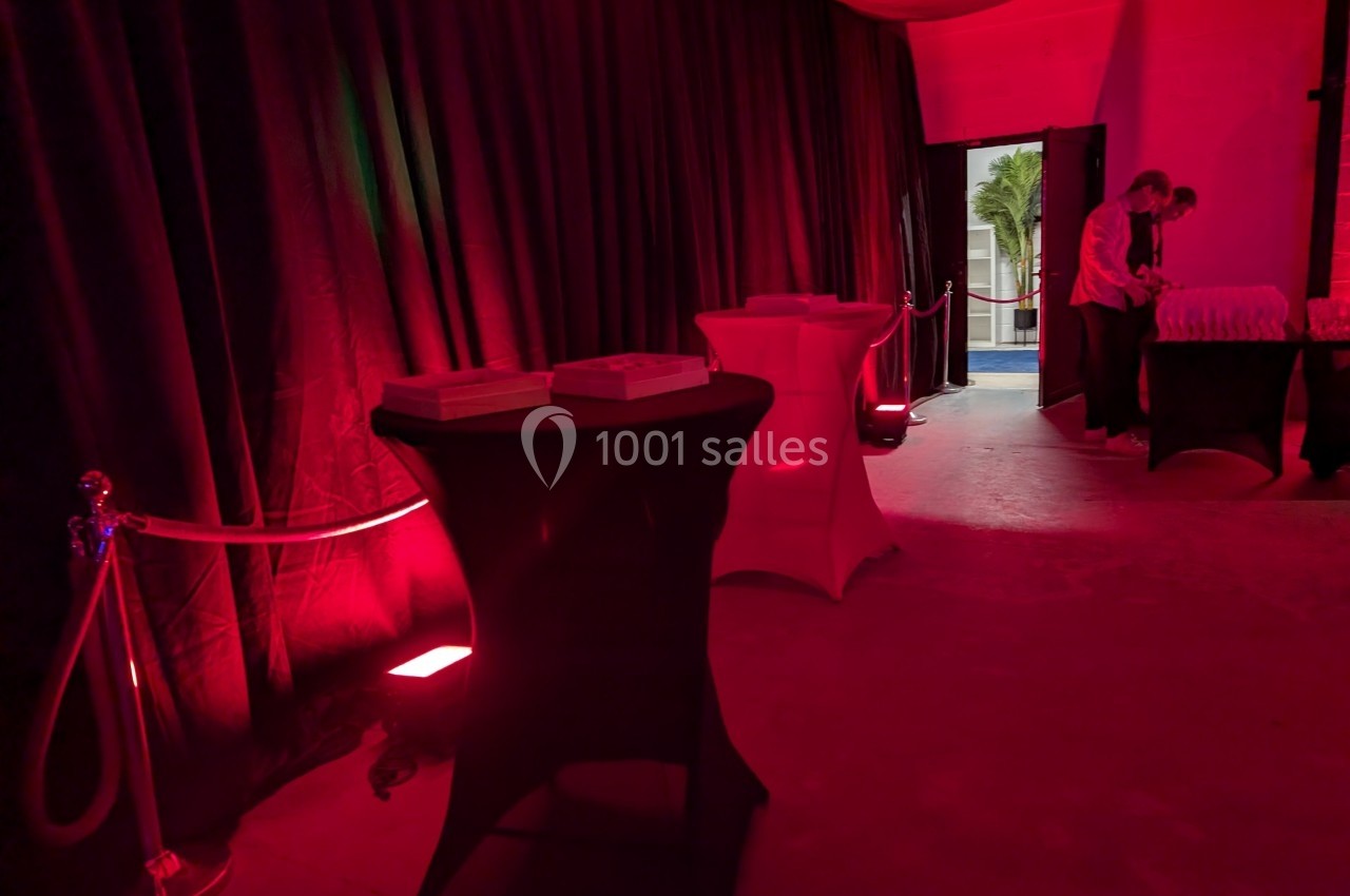 Salle faiblement éclairée avec des nappes rouges et noires sur des tables hautes, et deux personnes près d'une sortie.