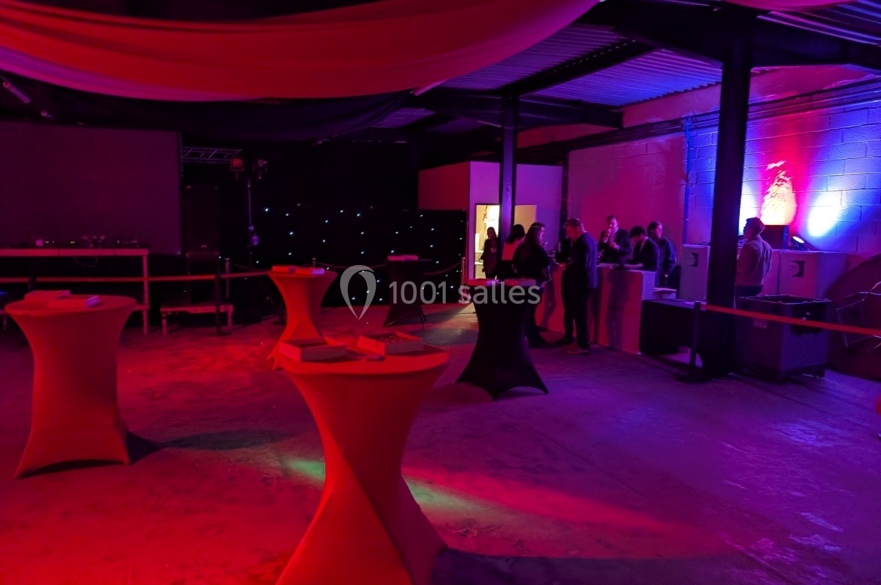 Salle sombre avec des tables hautes éclairées par des lumières rouges et bleues, et des personnes discutant près d'un…