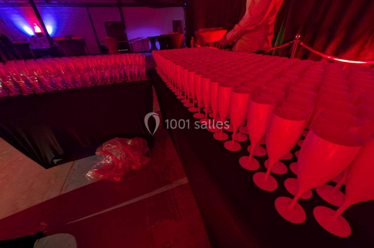 Rangées de verres à pied rouges alignés sur une table dans une salle éclairée par des lumières tamisées rouges et violettes.