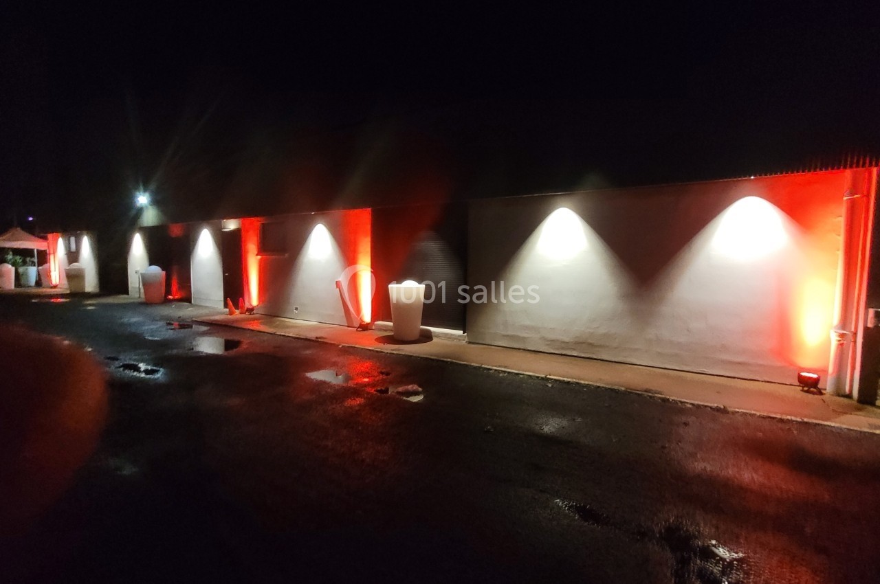 Façade d'un bâtiment éclairée par des lumières rouges et blanches, avec sol humide reflétant les éclairages.