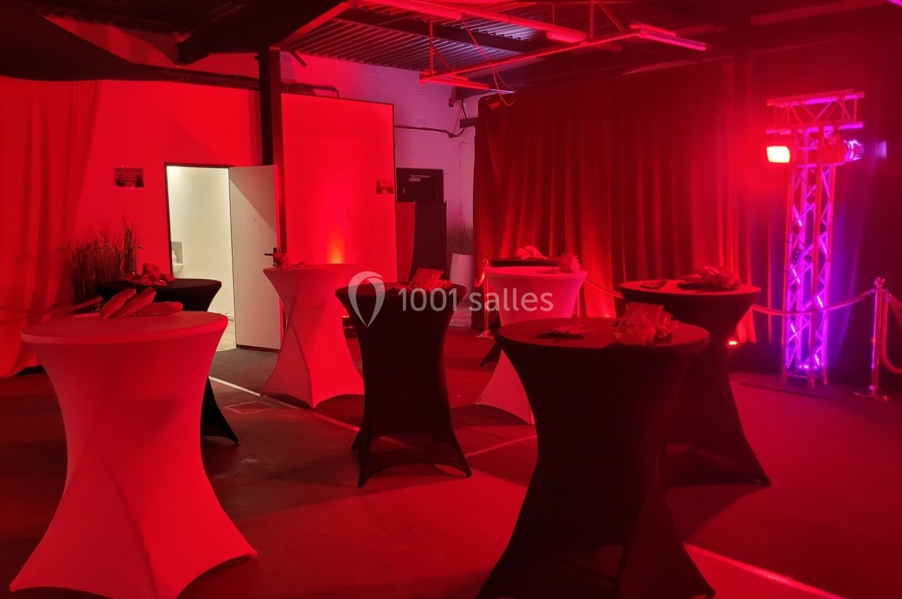 Salle sombre avec des tables hautes nappées de noir et blanc, éclairée par des lumières rouges et un projecteur.