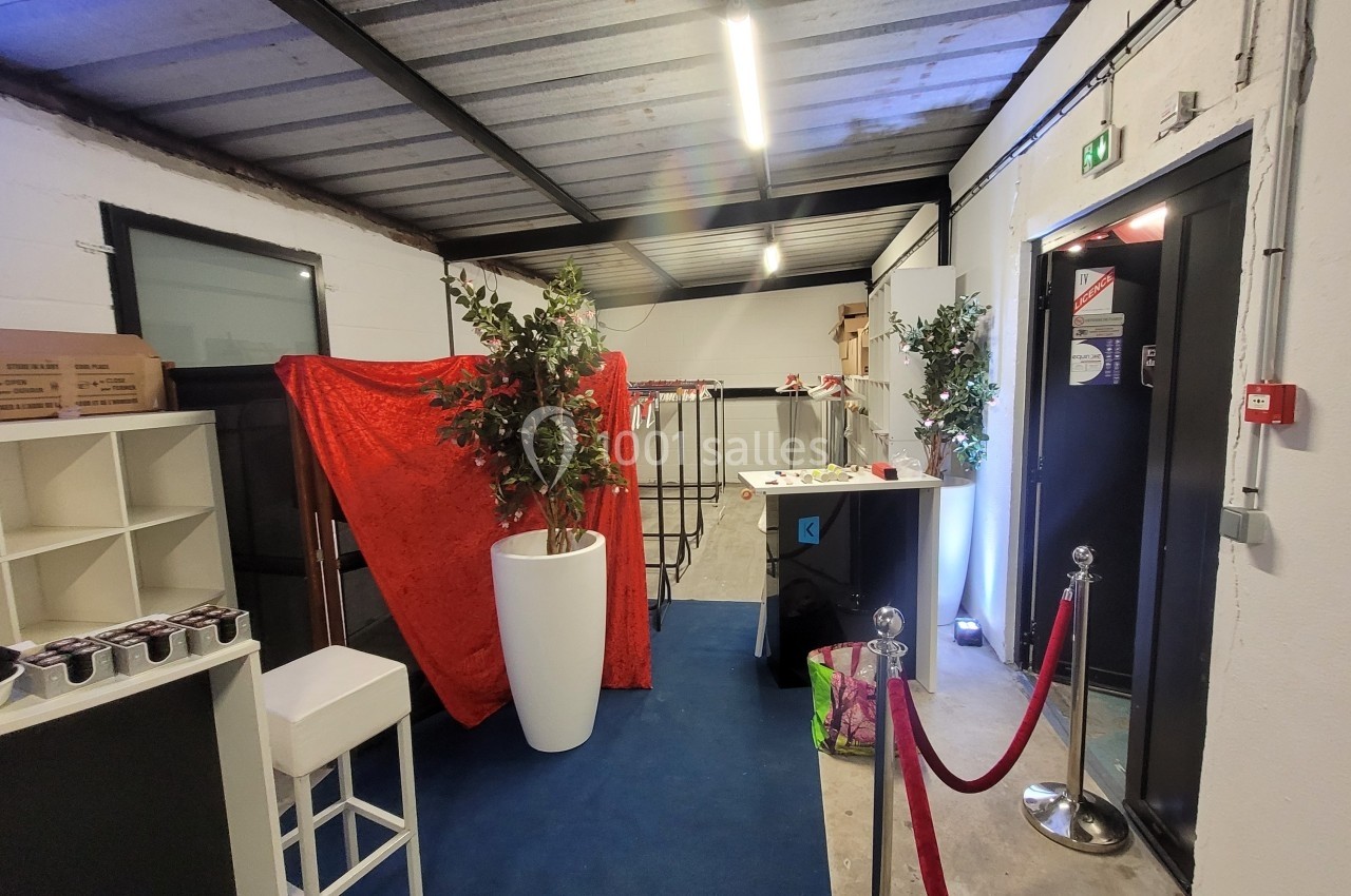 Espace intérieur avec sol bleu, plantes en pots, rideau rouge, mobilier blanc et noir, et barrière à cordon rouge.