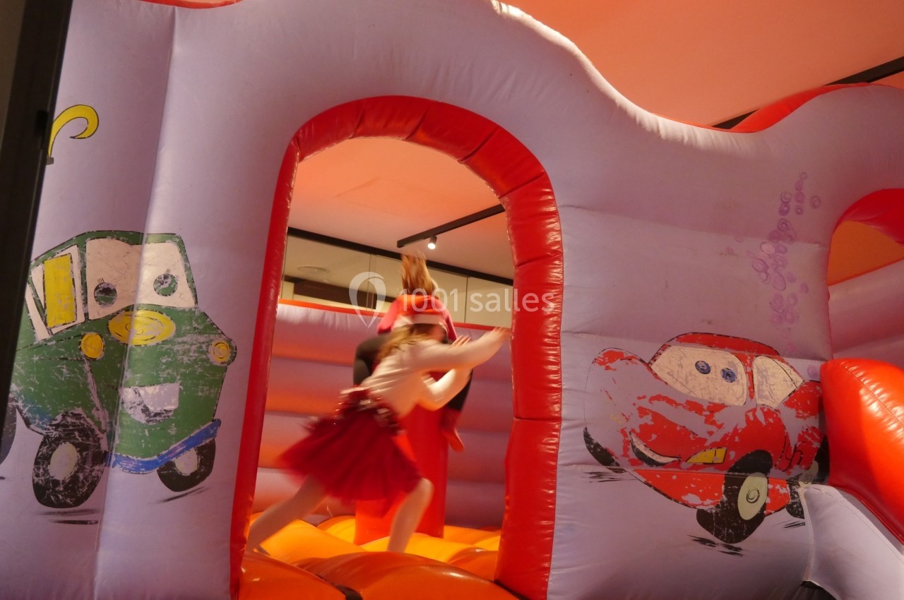 Une enfant en robe rouge joue sur un château gonflable décoré de voitures colorées dans une salle intérieure.