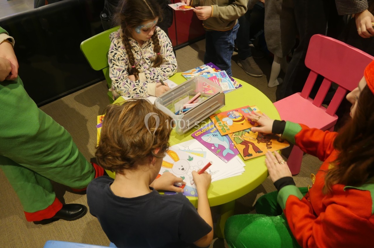 Des enfants colorient des dessins autour d'une table ronde, entourés d'adultes dans un espace intérieur animé.