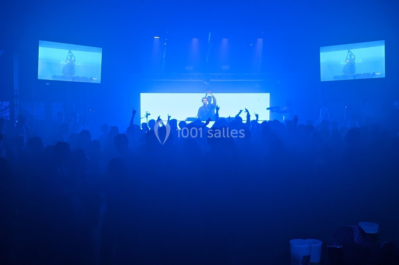 Foule dans une salle de concert sombre avec un DJ sur scène et des écrans lumineux en arrière-plan.