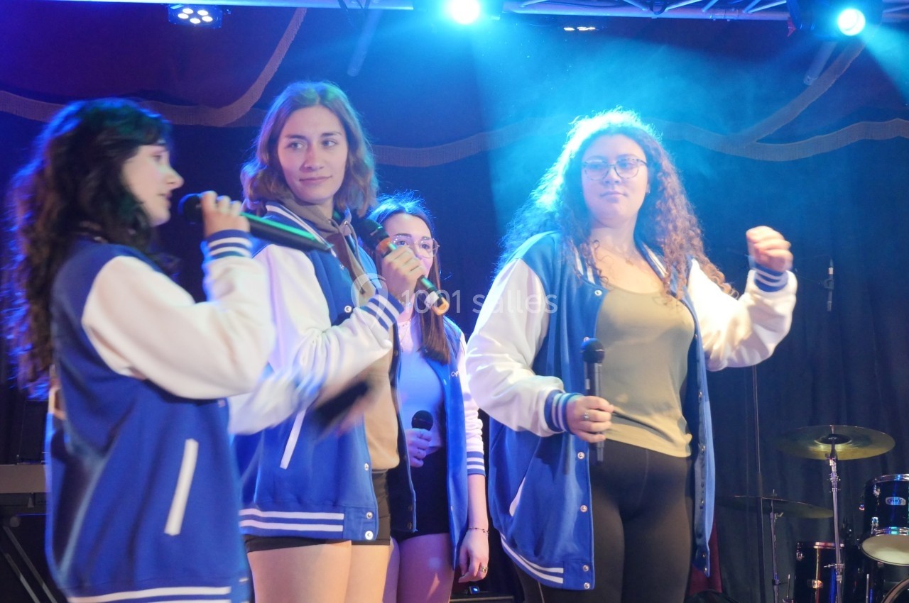 Quatre jeunes femmes en vestes bleues chantent sur scène, éclairées par des projecteurs, avec des micros en main.