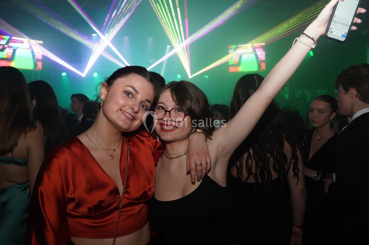 Deux femmes souriantes posent ensemble dans une ambiance festive avec des lumières colorées en arrière-plan.