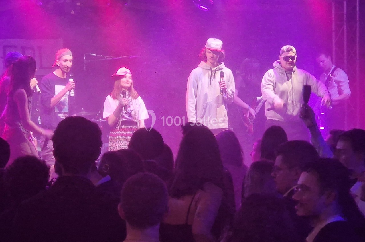 Groupe de chanteurs sur scène avec micros, éclairage rose et public debout dans une ambiance festive.