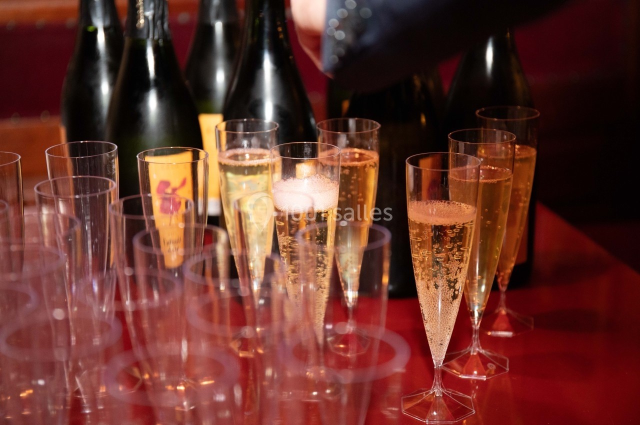 Flûtes de champagne remplies sur une table rouge, avec des bouteilles en arrière-plan.