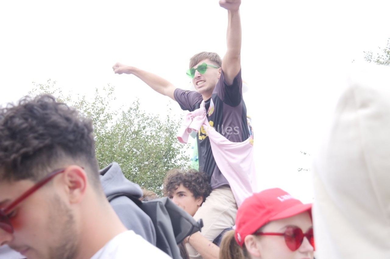 Un jeune homme portant des lunettes vertes lève les bras en l'air, assis sur les épaules d'une autre personne dans une foule.