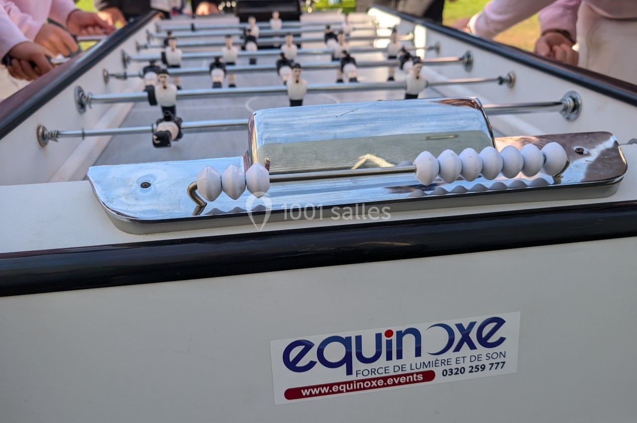 Vue rapprochée d'un baby-foot avec des joueurs miniatures blancs et noirs, entouré de personnes en arrière-plan.