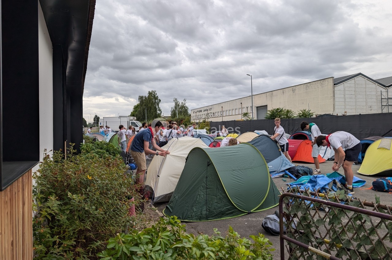 Des campeurs montent des tentes sur un terrain extérieur près de bâtiments industriels par temps nuageux.