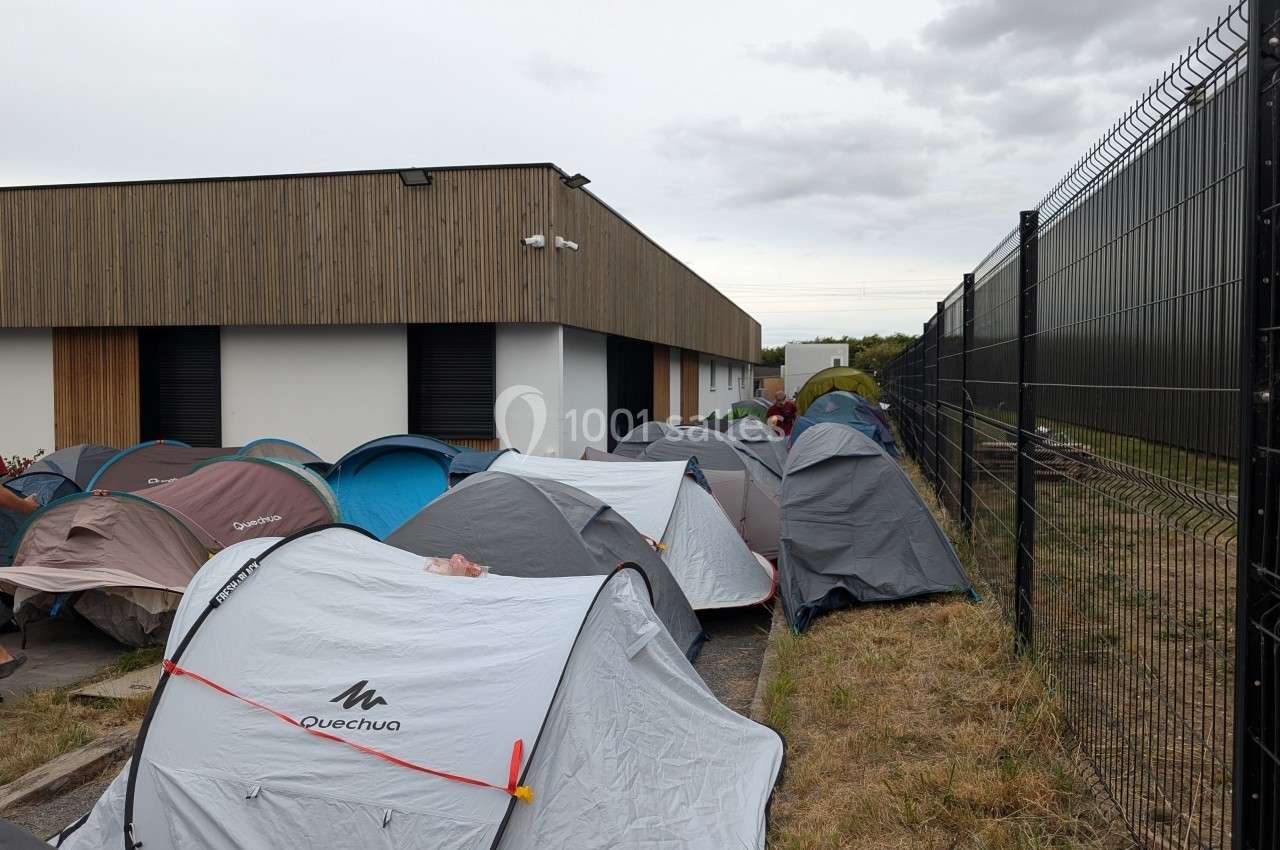 Des tentes alignées sur un sol herbeux près d'un bâtiment moderne et d'une clôture métallique sous un ciel nuageux.