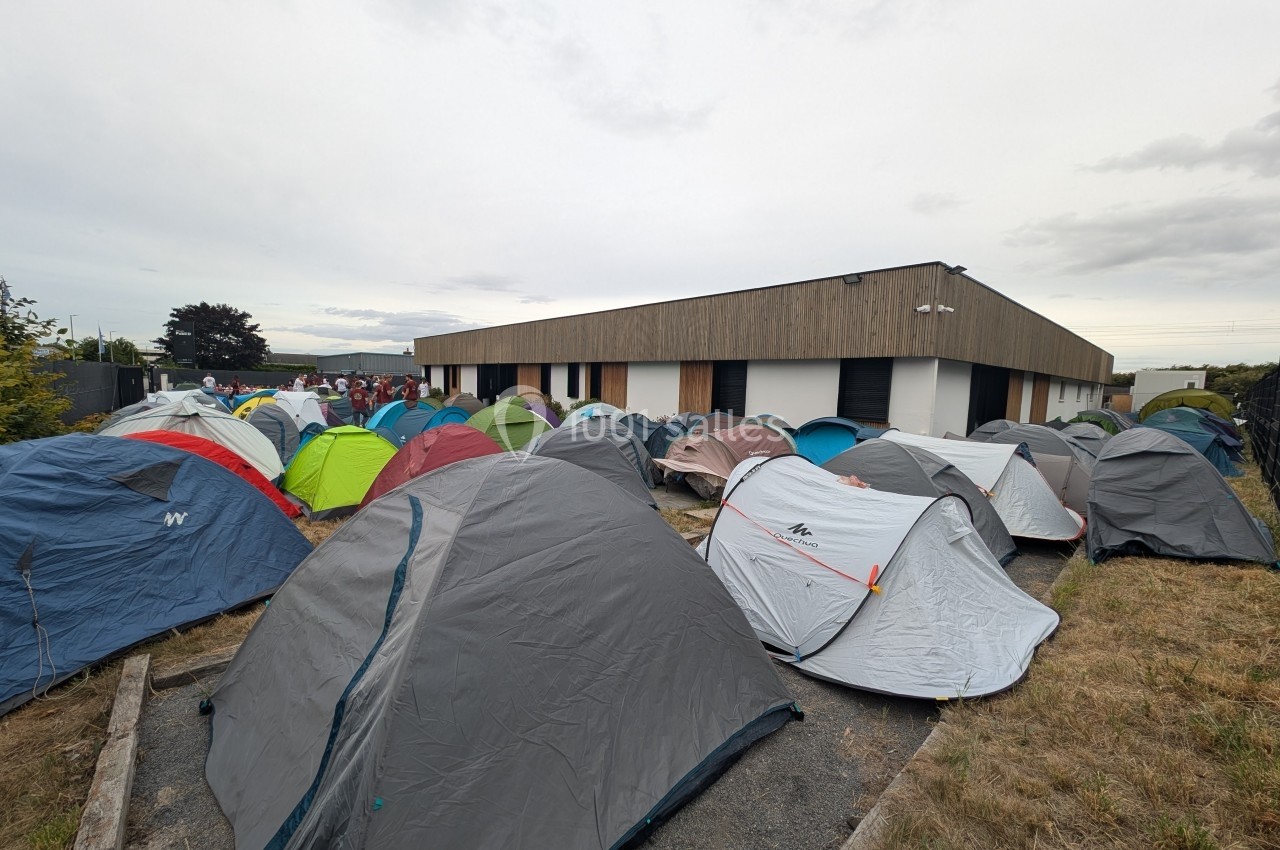 Alignement de tentes installées sur un terrain herbeux près d'un bâtiment moderne sous un ciel nuageux.