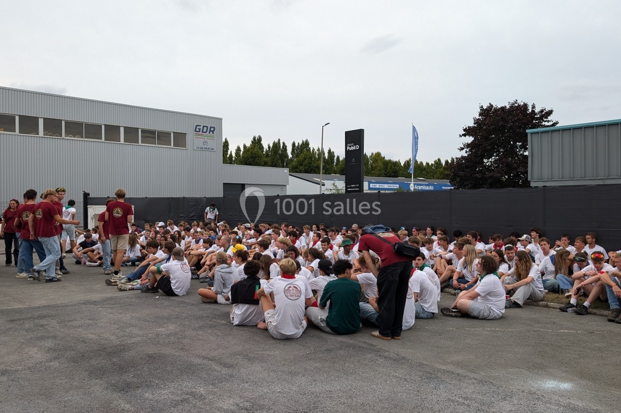 Un grand groupe de personnes assises sur un parking, certaines en t-shirts blancs, écoutant des intervenants debout.