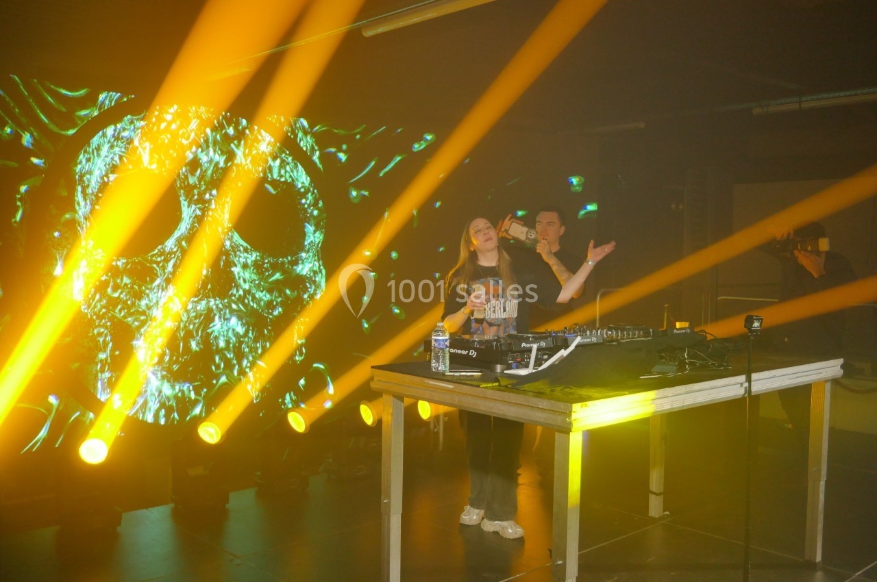 Deux personnes sur scène avec une table de mixage, éclairées par des projecteurs jaunes et un écran affichant un crâne…