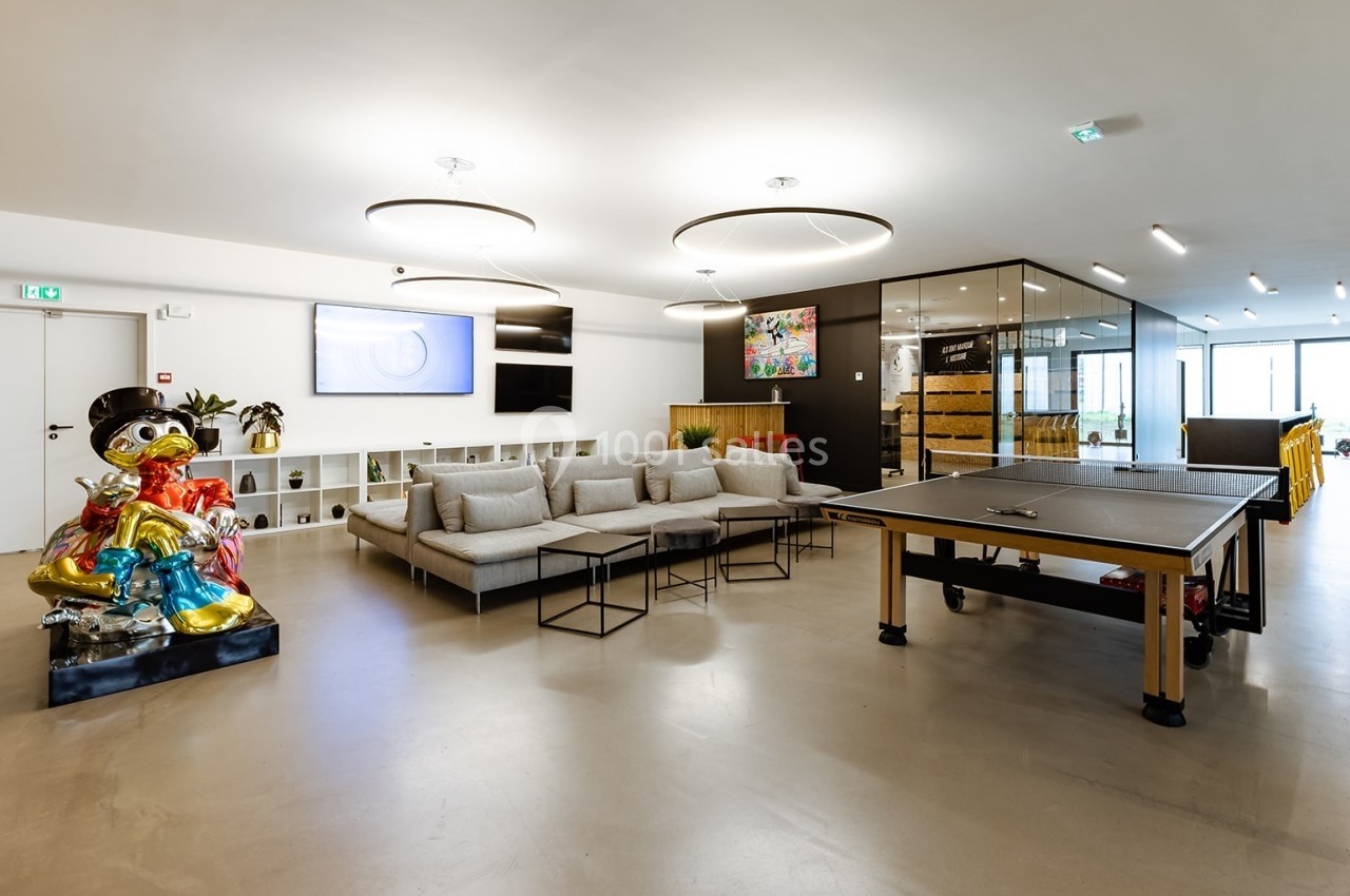 Espace intérieur moderne avec canapé, table de ping-pong, statue colorée et étagères, éclairé par des luminaires suspendus.