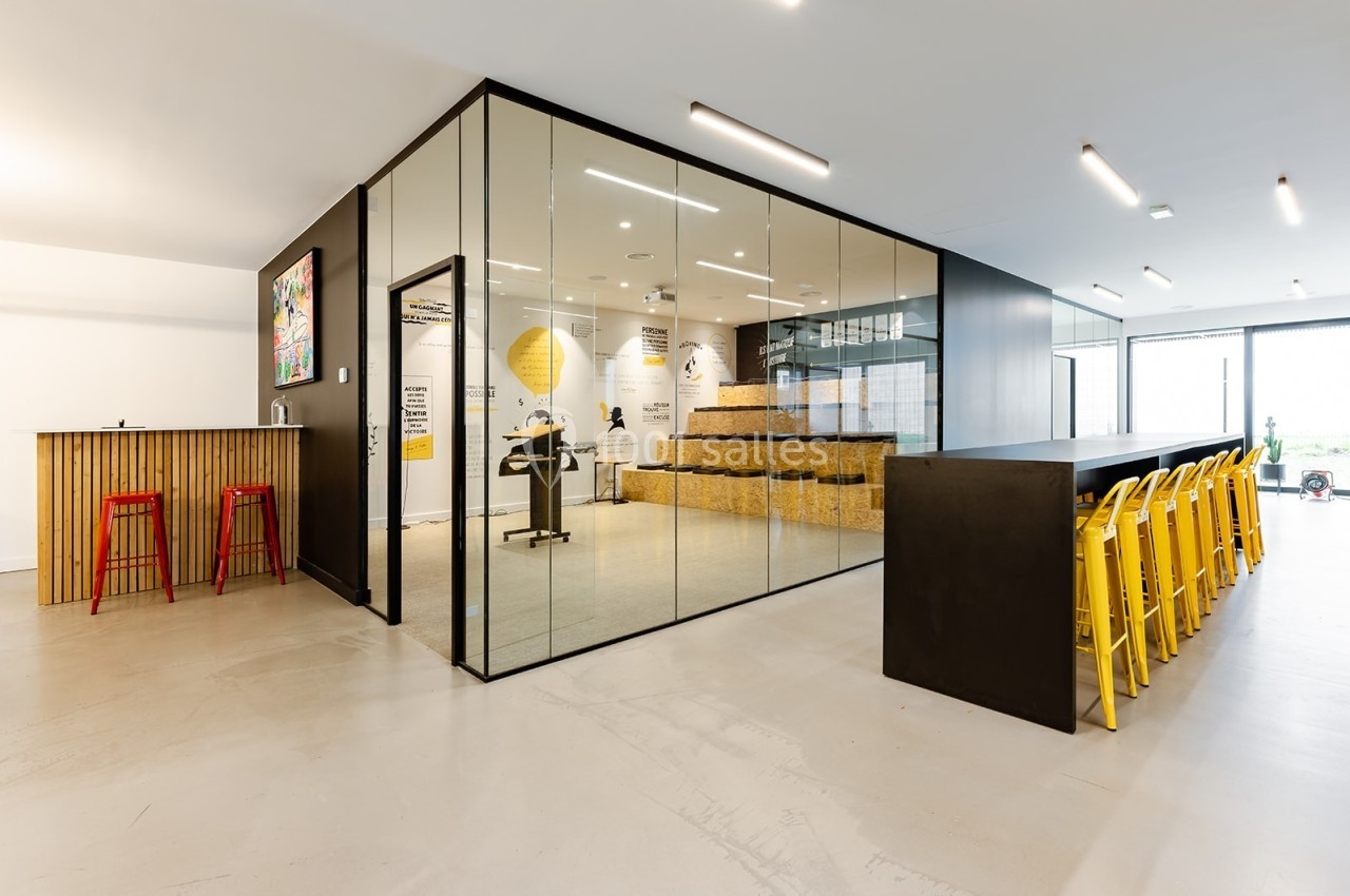 Salle moderne avec murs vitrés, espace de réunion en gradins, tabourets rouges et jaunes autour de tables noires.