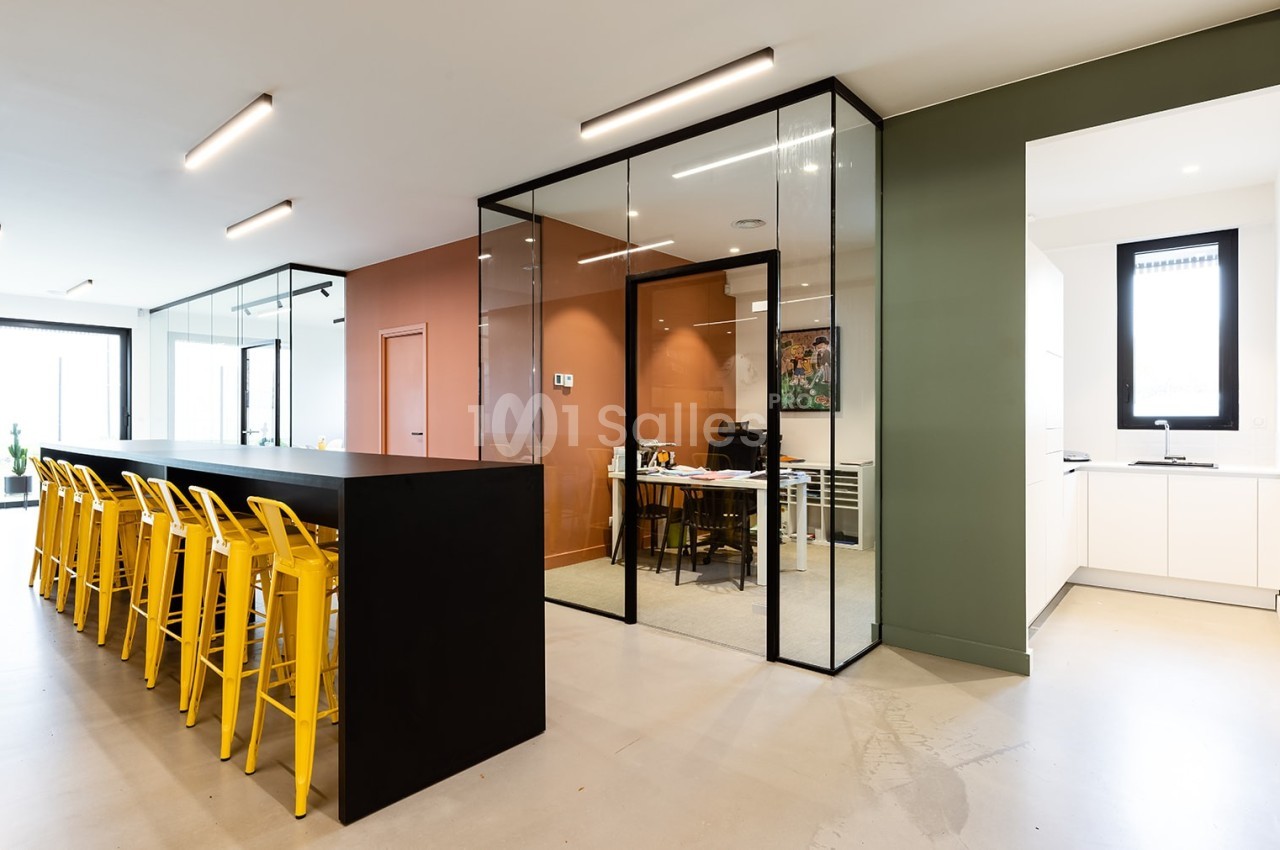 Espace de bureau moderne avec une salle vitrée, un long comptoir noir et des chaises jaunes alignées.