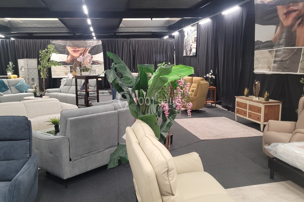 Salle d'exposition avec fauteuils, canapés, plantes décoratives et rideaux noirs en arrière-plan.