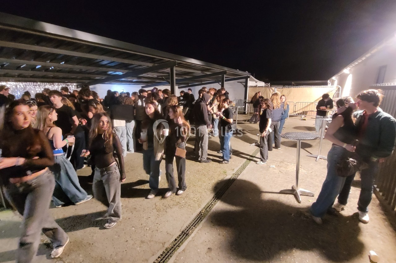 Groupe de personnes rassemblées en extérieur sous un abri, de nuit, certaines discutant ou tenant des boissons.