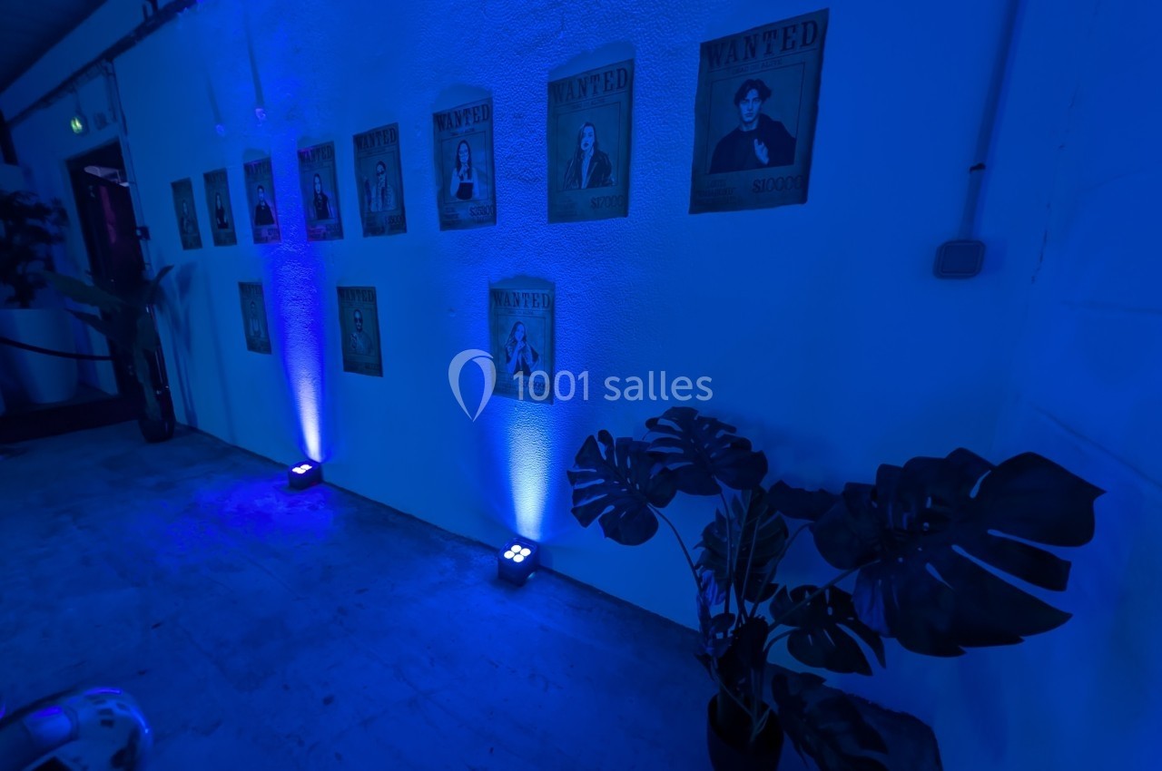 Posters ’Wanted’ éclairés en bleu accrochés sur un mur blanc, avec des plantes en pot au premier plan.