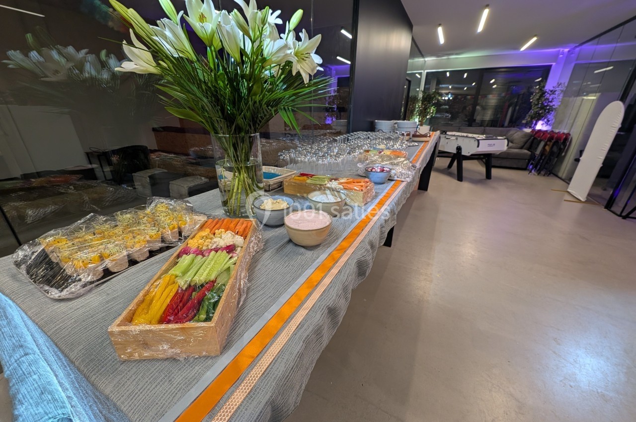 Table de buffet avec fleurs blanches, légumes, sauces, desserts et verres, dans un espace moderne et éclairé.