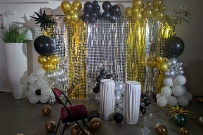 Miniature Location salle Seclin (Nord) - Luminaria #284 Décor festif avec ballons noirs, dorés, argentés et blancs, fond scintillant et deux tabourets blancs devant.