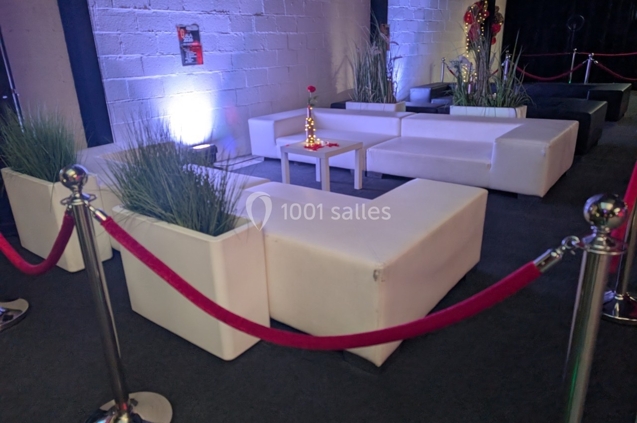 Espace lounge avec canapés blancs, table basse et plantes décoratives, délimité par des cordons rouges.