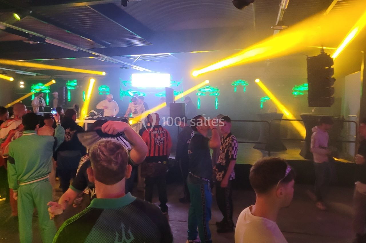 Personnes dans une salle de concert avec des jeux de lumières colorées et des DJ sur une scène en arrière-plan.