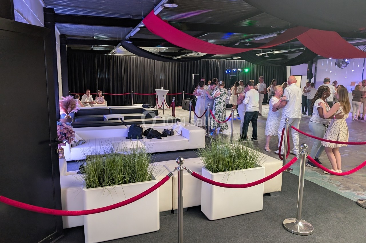 Salle de réception avec canapés blancs, plantes décoratives et invités debout ou discutant près des tables.